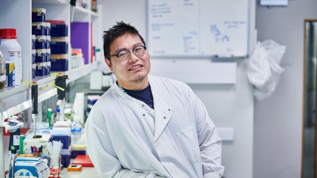 A smiling scientist in a white coat leans against a lab bench