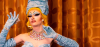 In this image, SALLY is pictured posing towards camera. The picture is framed from head to just below their shoulders. They are a drag artist, with bright blonde hair down to their shoulders. They are wearing pale blue crinkled gloves up to their biceps, with a matching head piece. They are also wearing a jewel necklace, with 6 rows of different jewels. They have hoop gold earrings that read 'babygirl' inside. 